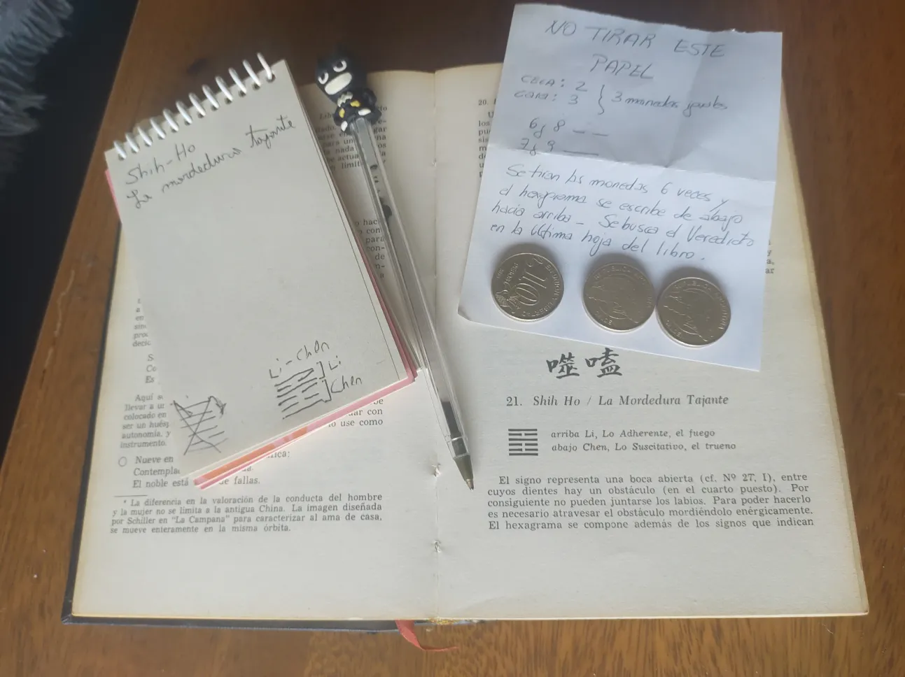 Various items related to Yijing. The Book of Changes is open on the page containing the description for Gnawing Bite. Two pieces of paper are laid on top of the book, one containg two trigrams, and the other containing instructions on how to toss Yijing. Three coins can be seen.